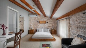 A bedroom with a stone wall, wooden beams, a bed, a desk, a chair, and a nightstand with a lamp. - Hotel La Grisa (Bale)