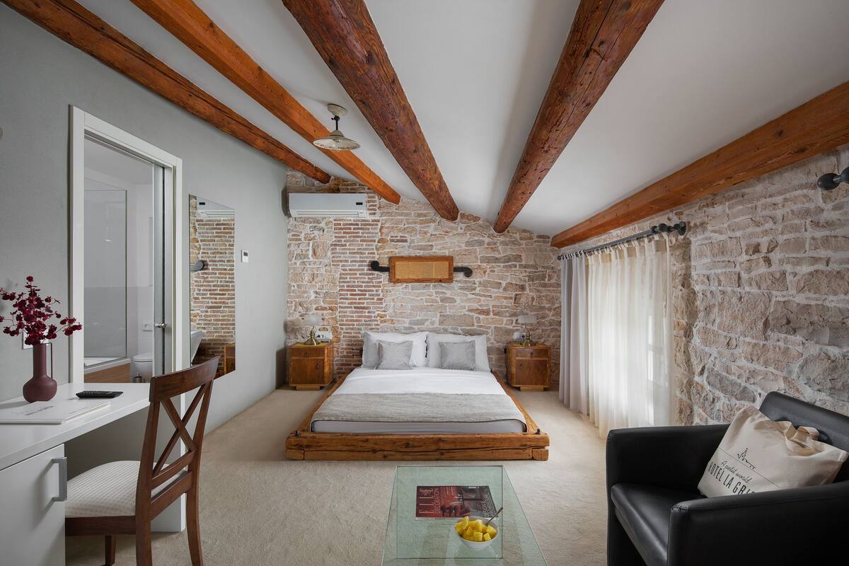 A bedroom with a stone wall, wooden beams, a bed, a desk, a chair, and a nightstand with a lamp.