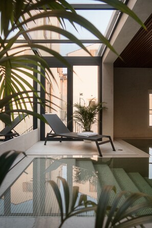 A modern indoor pool with clear glass walls, lounge chairs, and potted plants. - Hotel La Grisa (Bale)