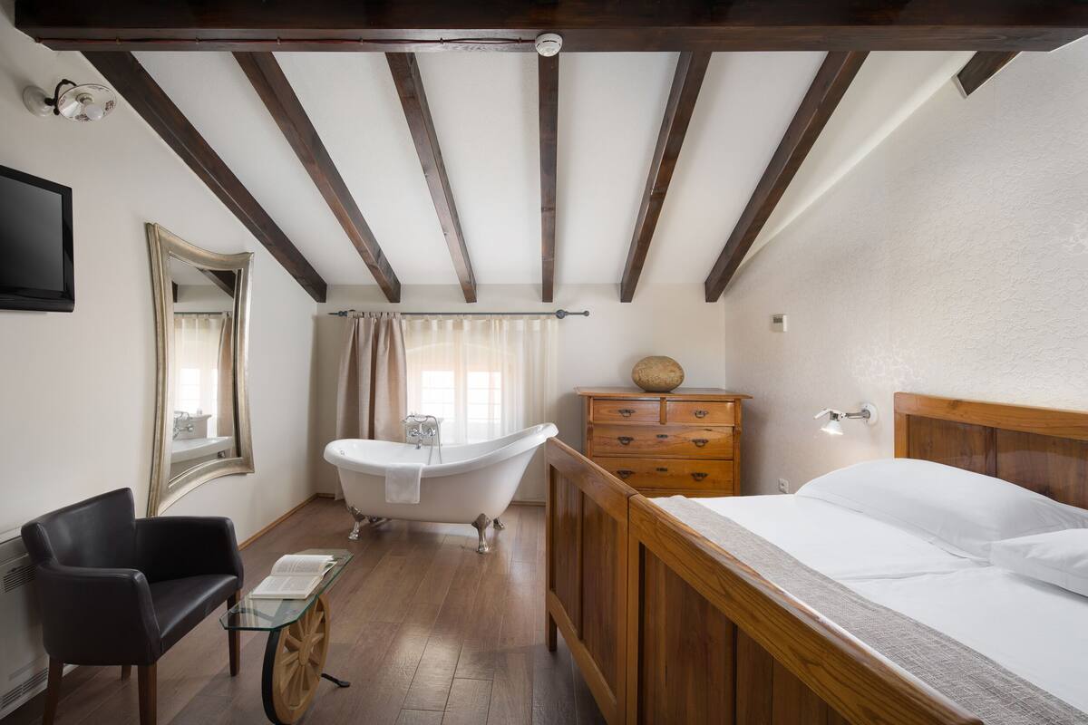 A room with a wooden bed, a black chair, a glass table, a white bathtub, and a wooden dresser.