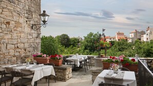 An outdoor dining area with stone walls, tables with white tablecloths, and potted flowers. - Hotel La Grisa (Bale)