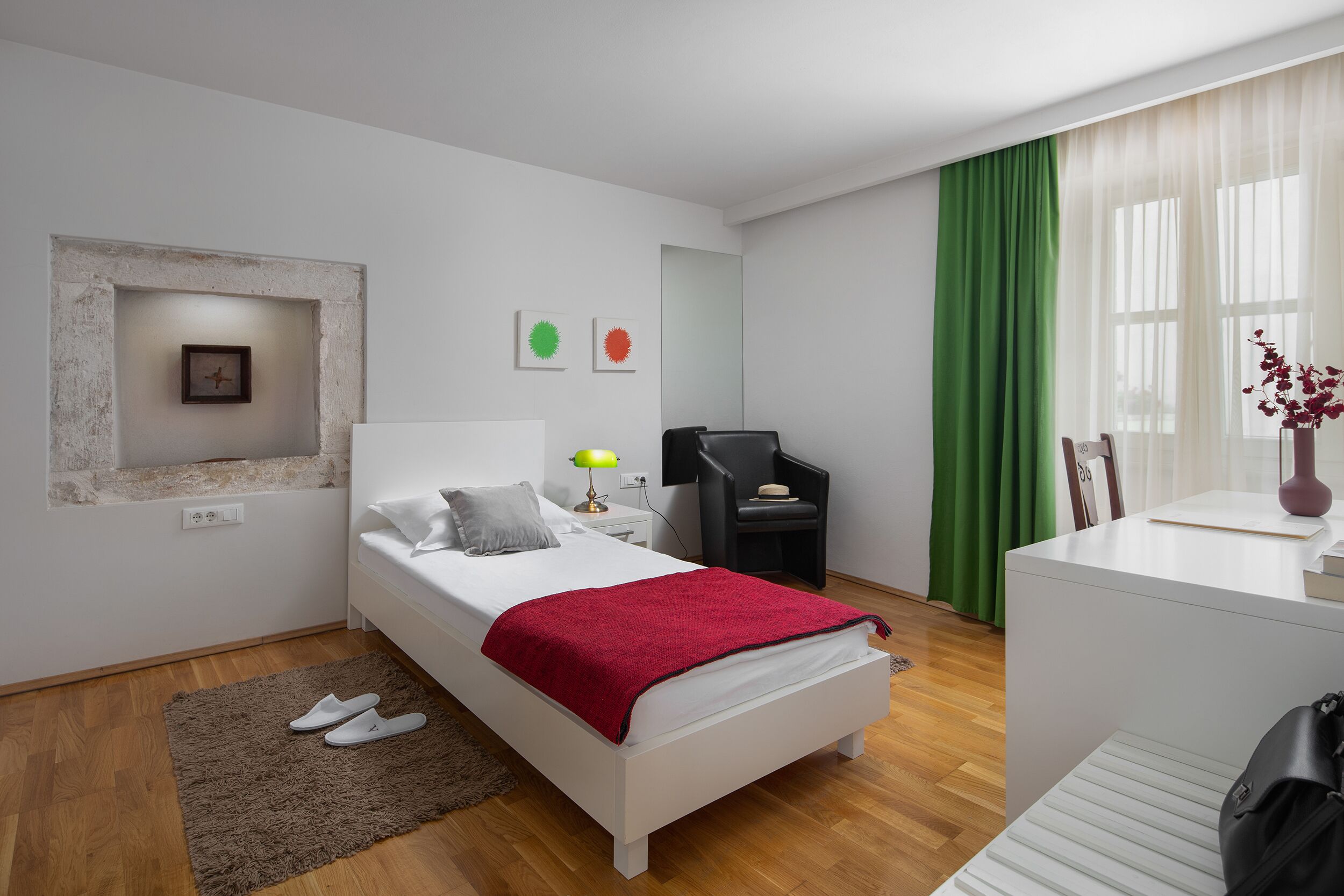 A modern bedroom with a white bed, a red throw blanket, a small desk, a chair, and a green curtain.