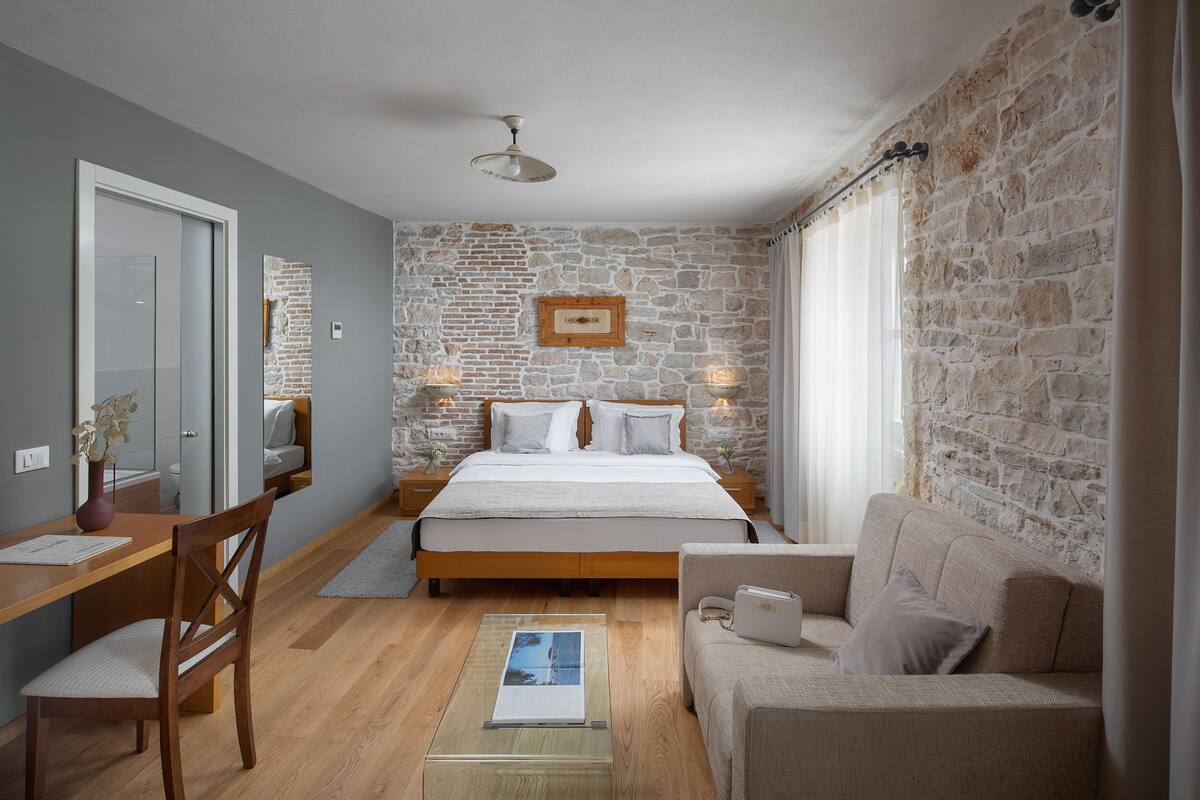 A bedroom with a stone wall, a bed, a desk, a chair, and a sofa.