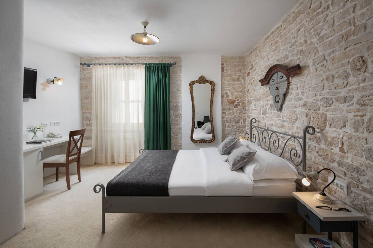 A bedroom with a stone wall, a bed with white bedding, a desk with a chair, a mirror, and a lamp.