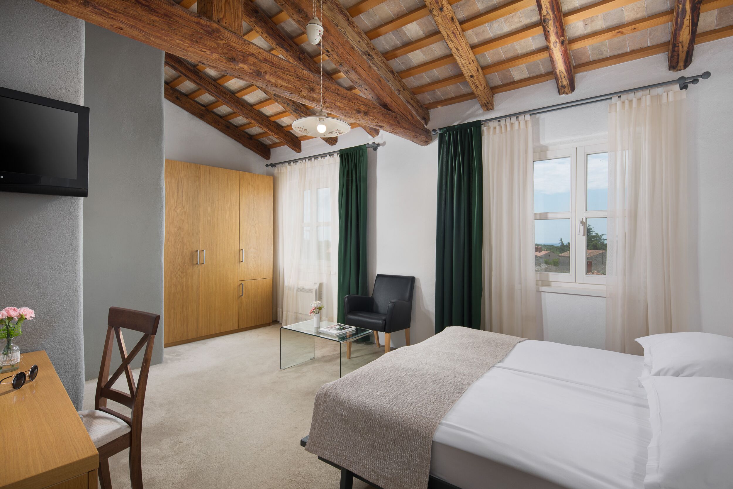 A hotel room with a bed, a desk with a chair, a TV, a wooden wardrobe, and a window with curtains.