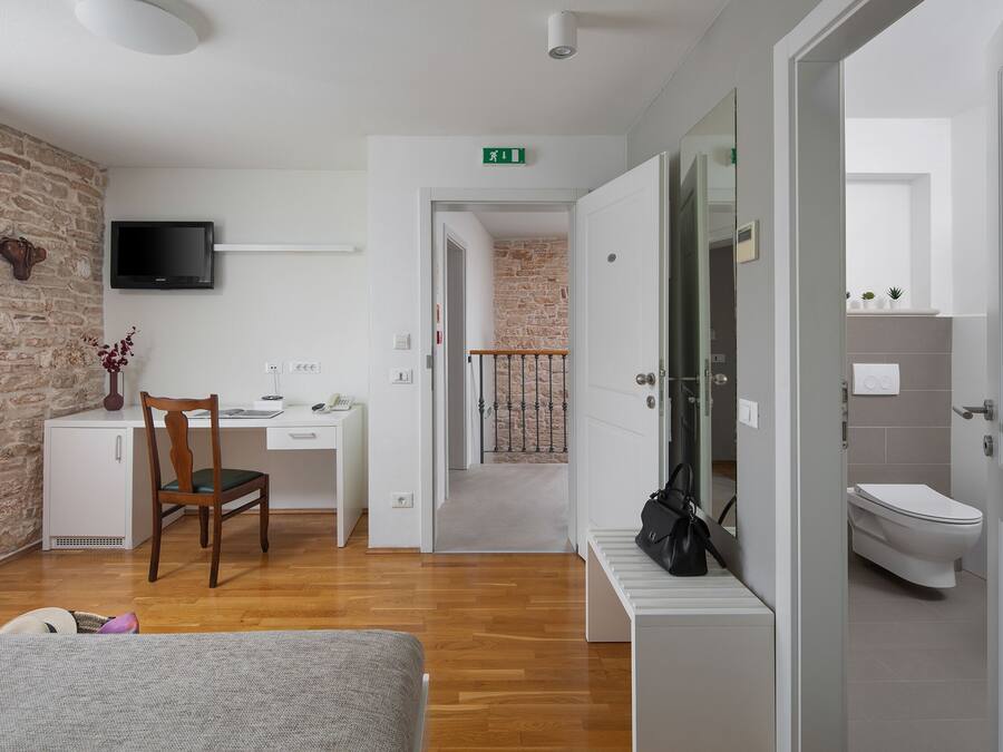 A hotel room with a wooden floor, a desk with a chair, a television mounted on the wall, and a bathroom visible through an open door.