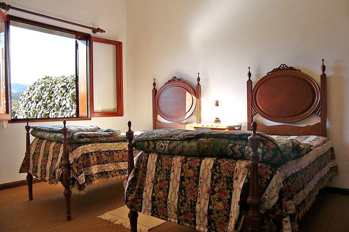 Quarto com duas camas de madeira, uma janela com vista e uma mesa de cabeceira com abajur.