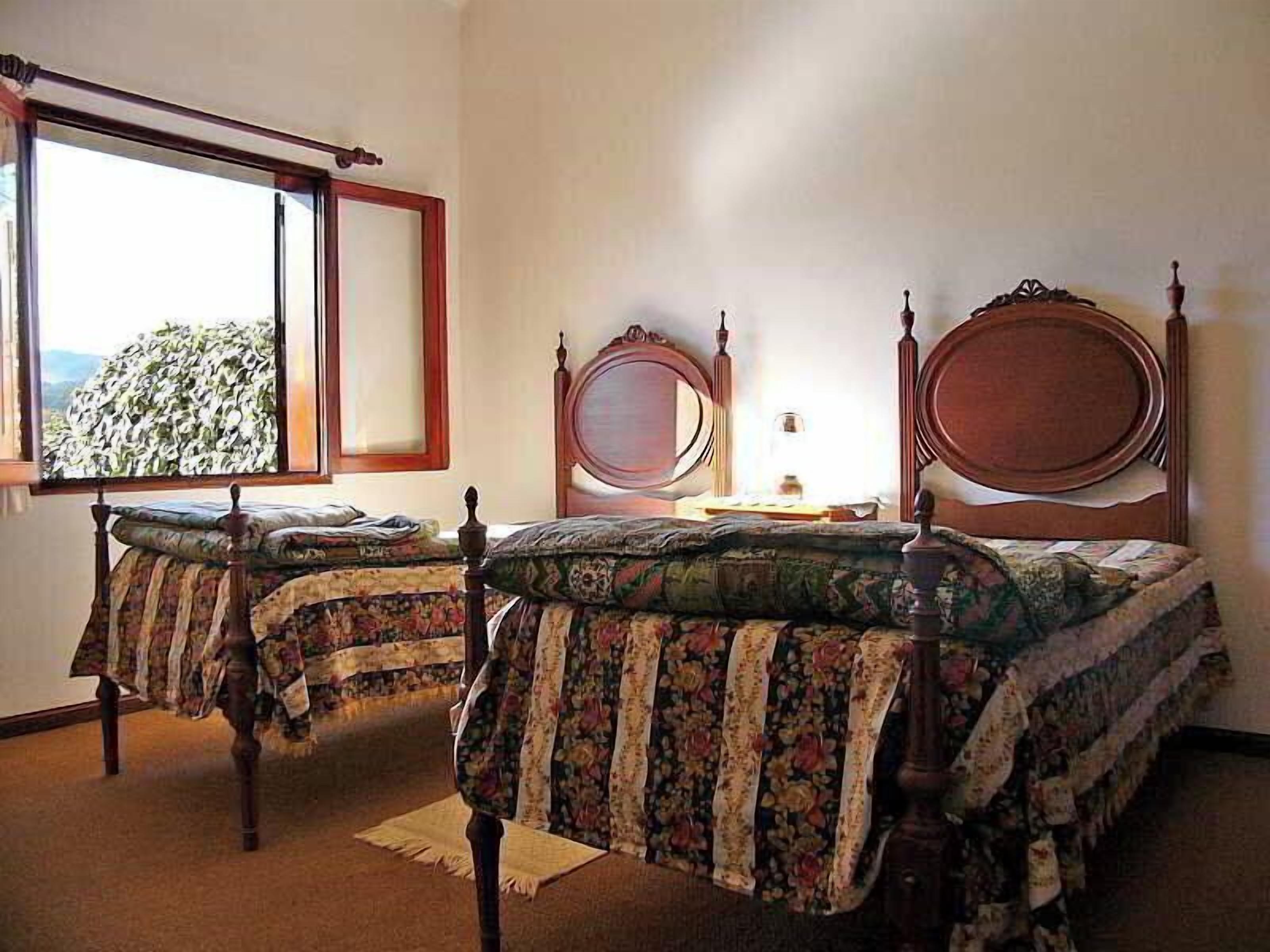 A room with two wooden beds with floral bedding, a window with a view, and a bedside table with a lamp.