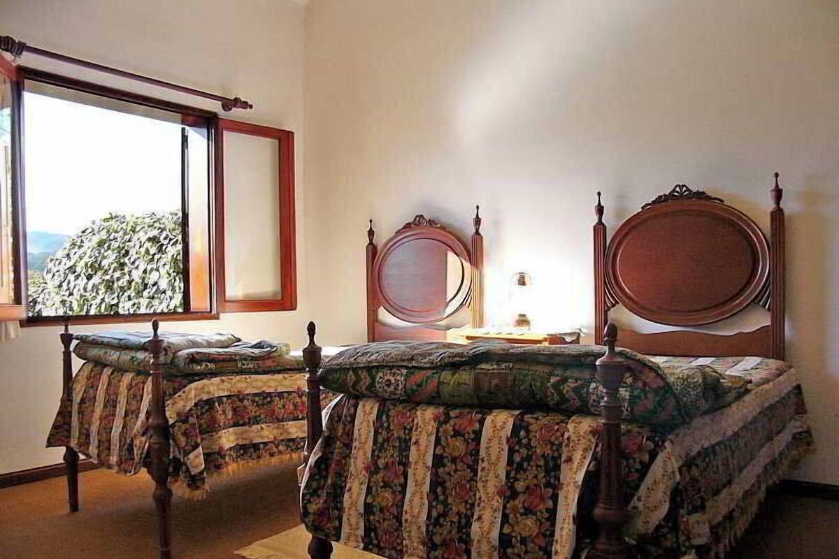 Quarto com duas camas de madeira com roupa de cama floral, uma janela com vista e uma mesa de cabeceira com abajur.