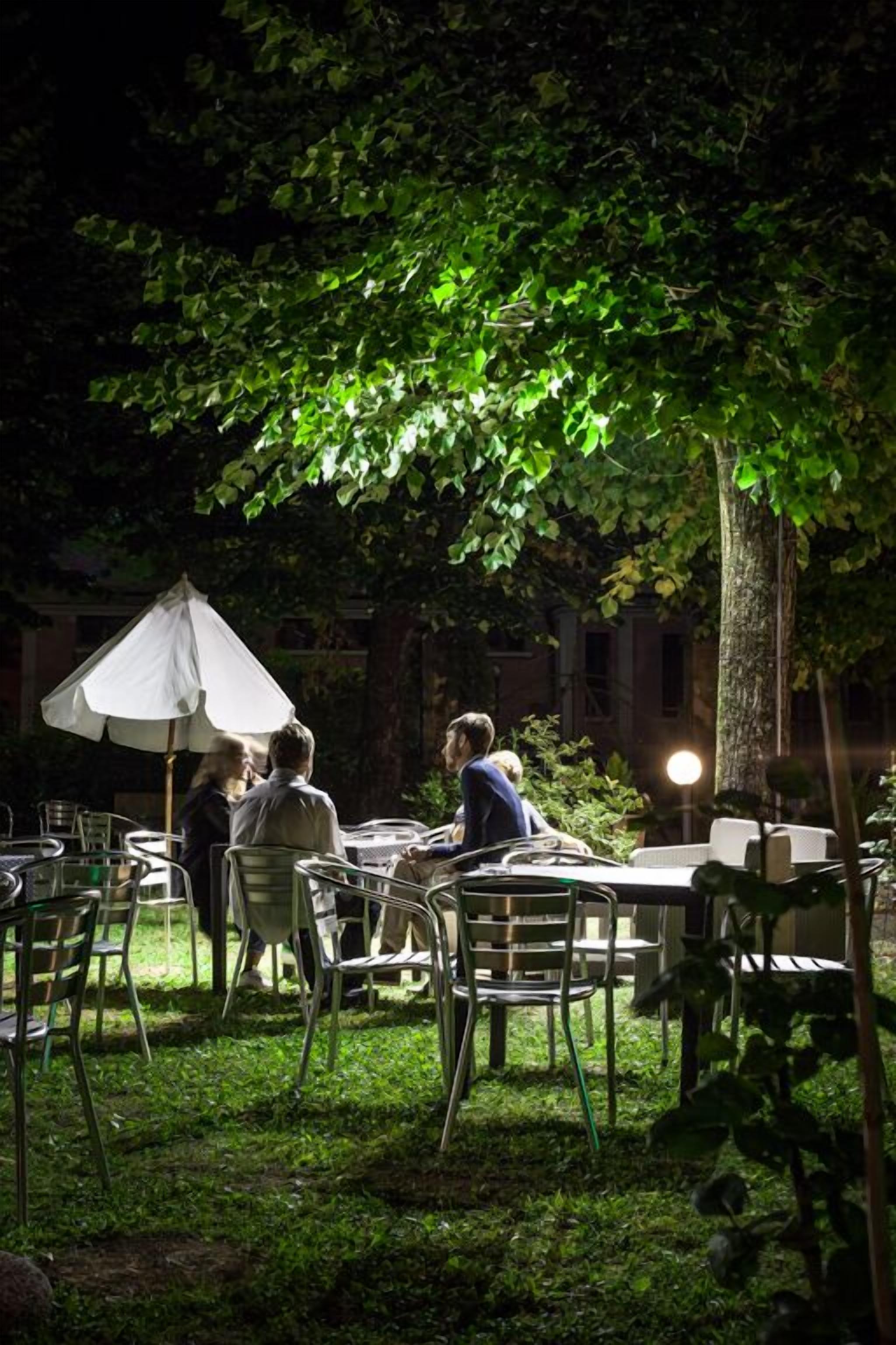 Un área de comedor al aire libre con sillas y mesas de metal, rodeada de follaje y árboles.