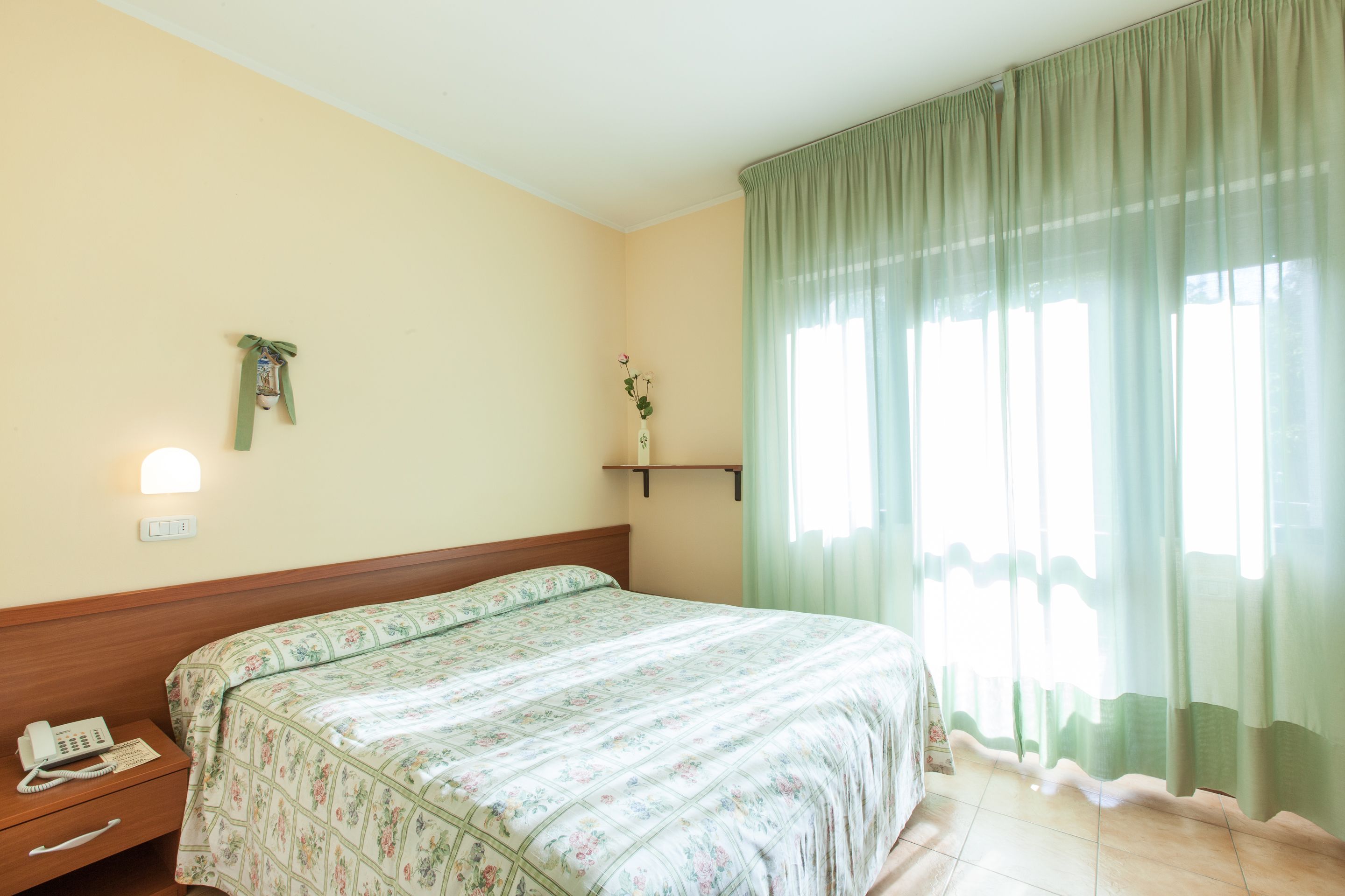 A hotel room with a bed, a bedside phone, a wall-mounted light, a small shelf with a vase, and a window with sheer curtains.