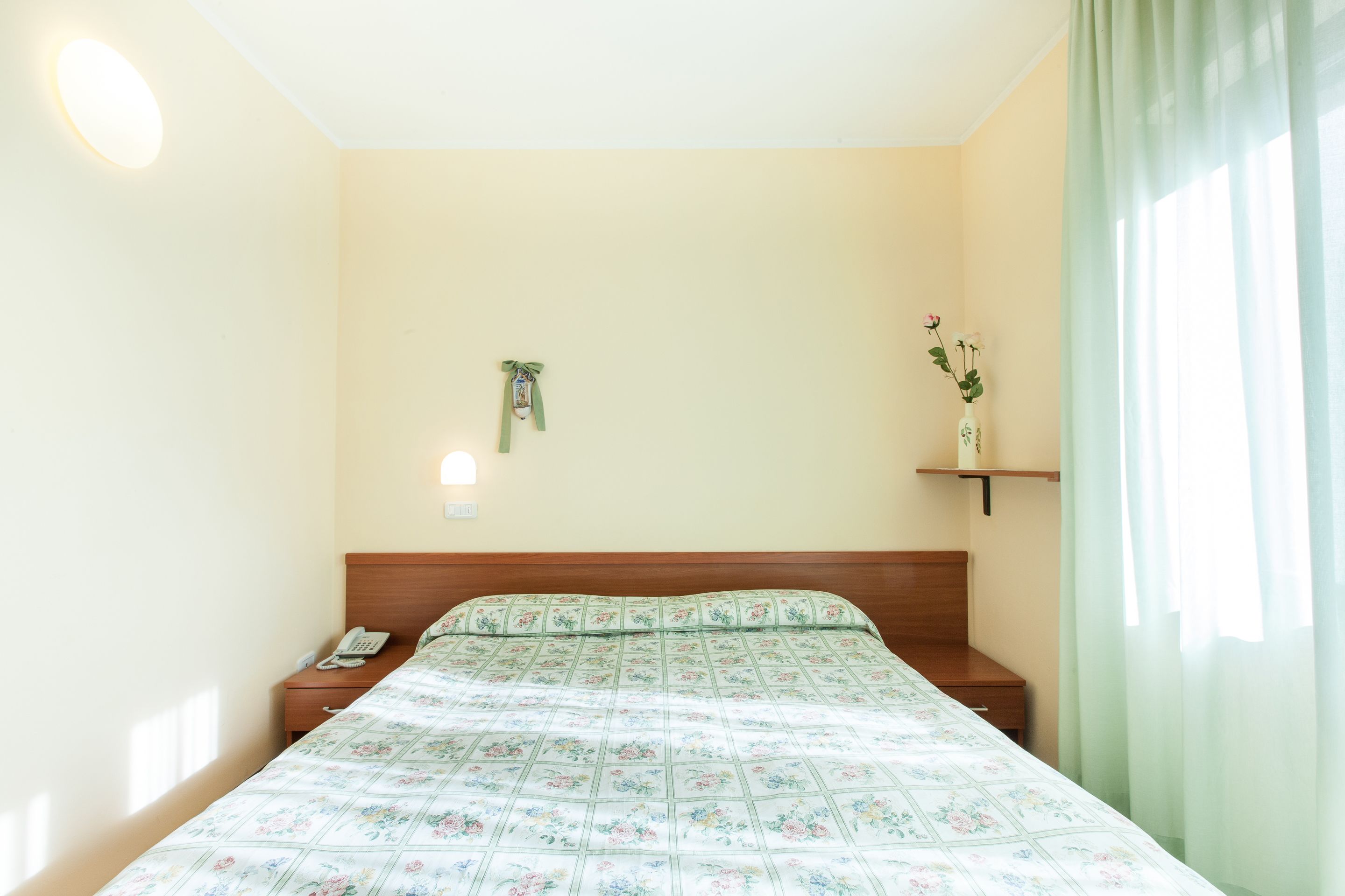 A hotel room with a bed, bedside tables, a telephone, a wall-mounted light, a shelf with a vase, and a window with curtains.