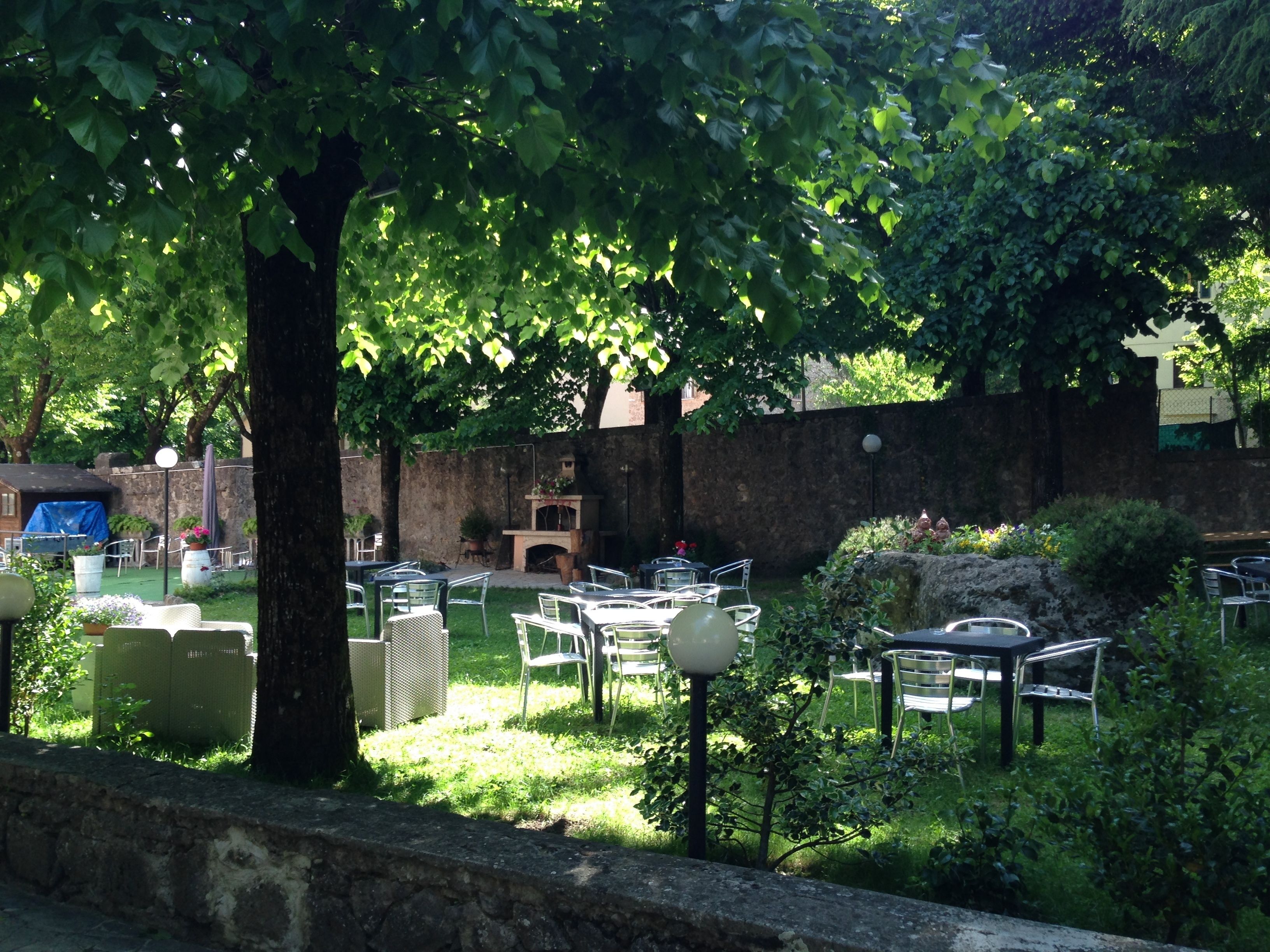 An outdoor seating area with tables and chairs, surrounded by greenery and a stone wall.