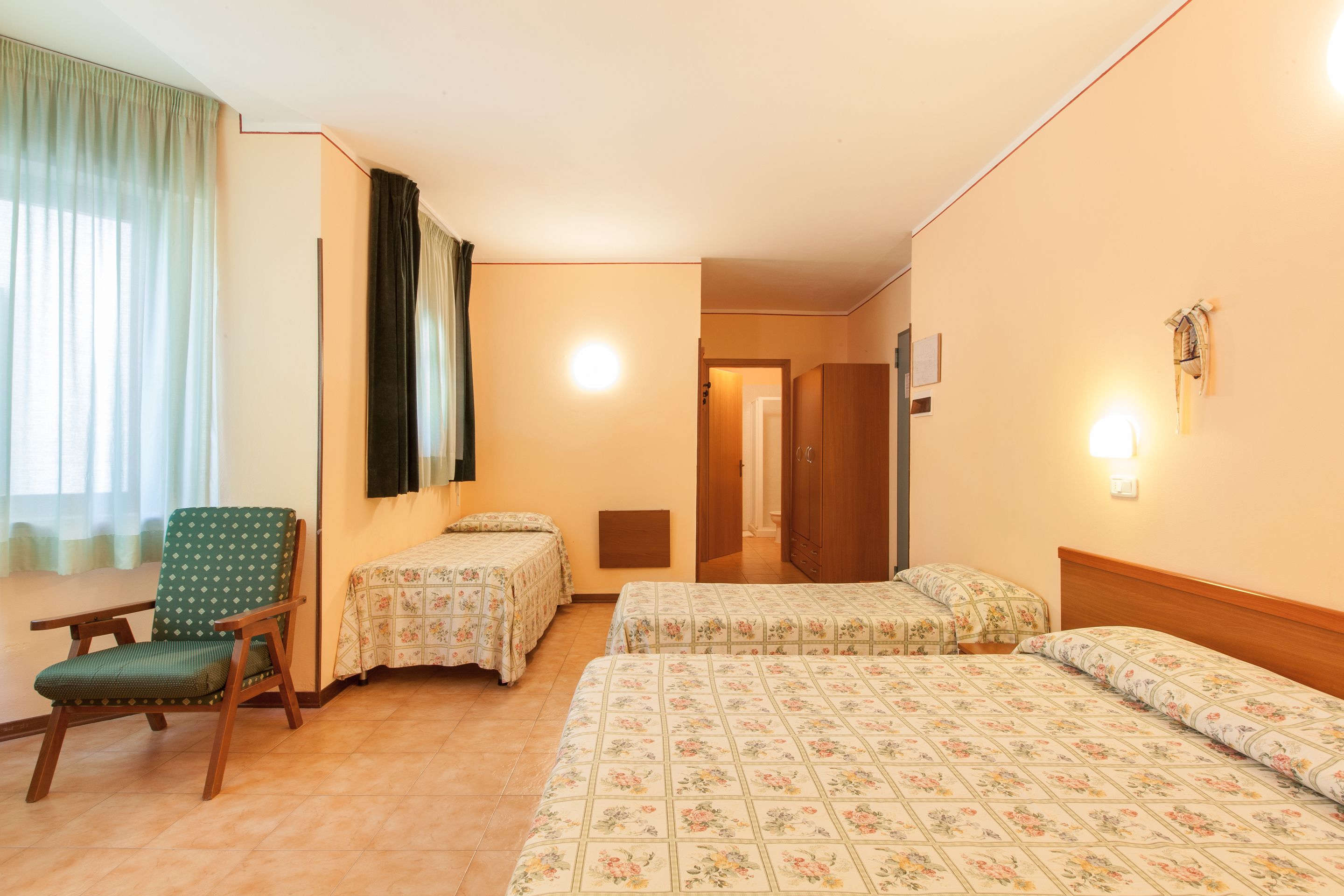 Habitación de hotel con dos camas individuales, una silla, un escritorio y vistas a un pasillo.