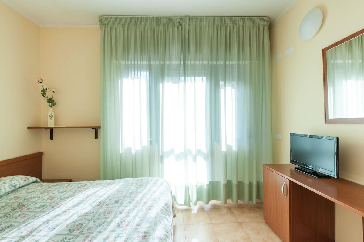 Une chambre à coucher avec un lit, un téléviseur posé sur un meuble en bois et une fenêtre ornée de rideaux légers.