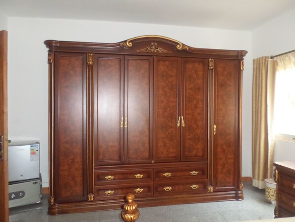 Un armario grande de madera con varios cajones y puertas, ubicado en una habitación que tiene un refrigerador y una ventana con cortinas.