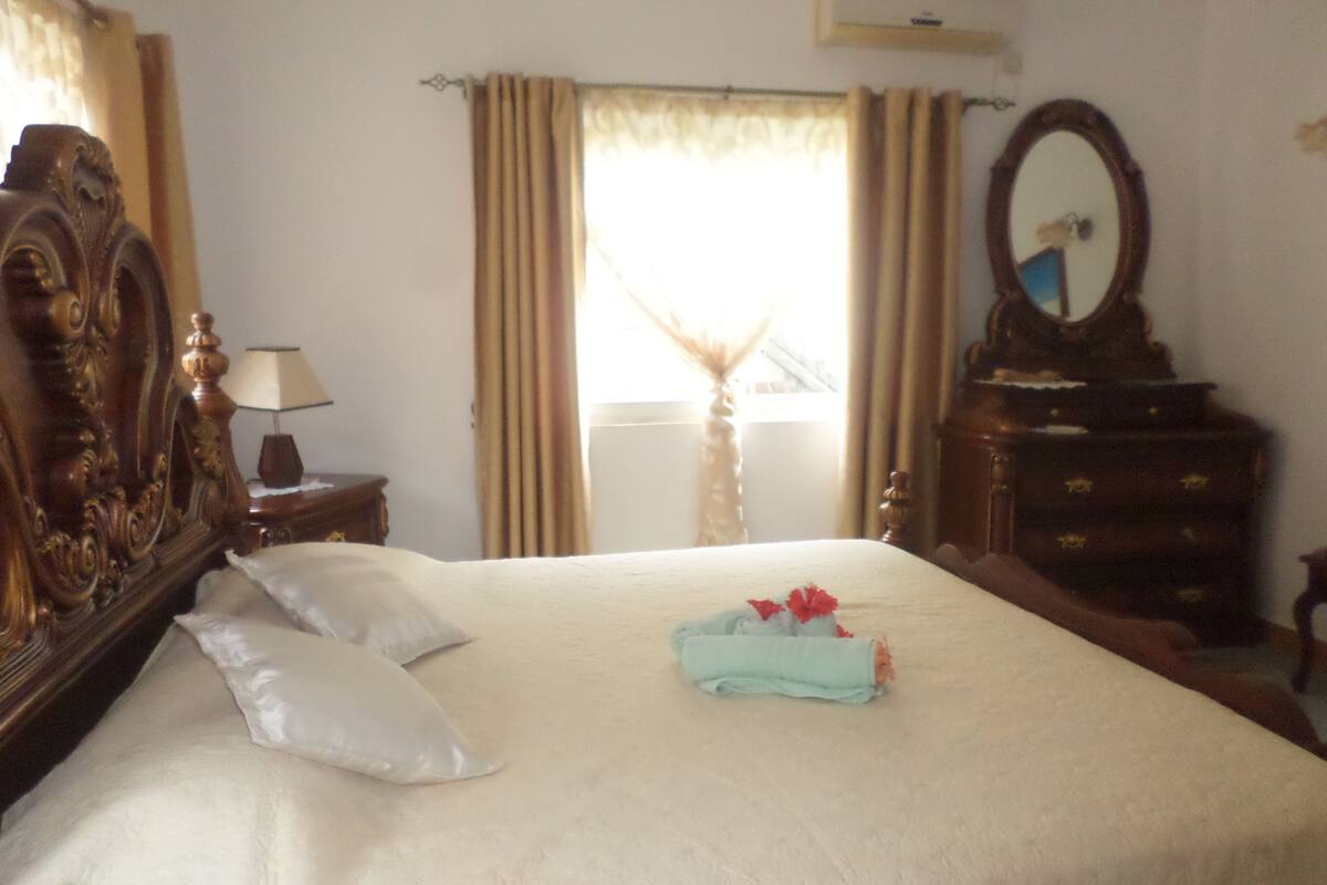 Una cama bien hecha con almohadas y una toalla doblada, un aparador de madera, un espejo y una lámpara en una mesita de noche.