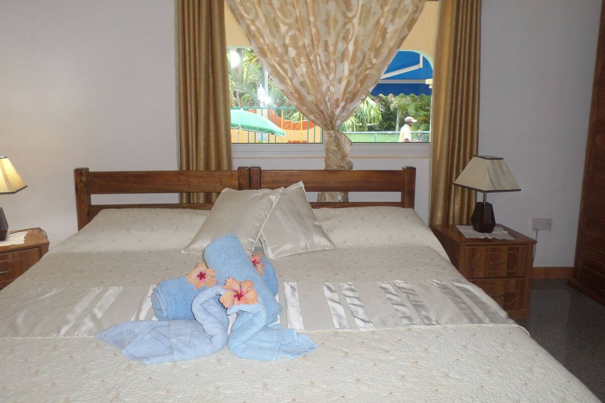 Una cama bien hecha con toallas y flores, un cabecero de madera, una mesita de noche con lámpara y una ventana con cortinas.