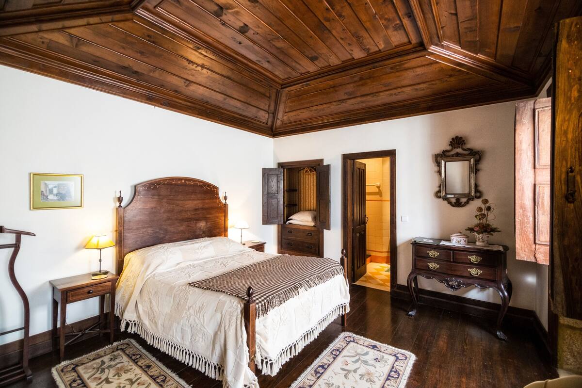 A traditional bedroom with a wooden ceiling, a large bed with a headboard, a nightstand, a dresser, and a mirror.