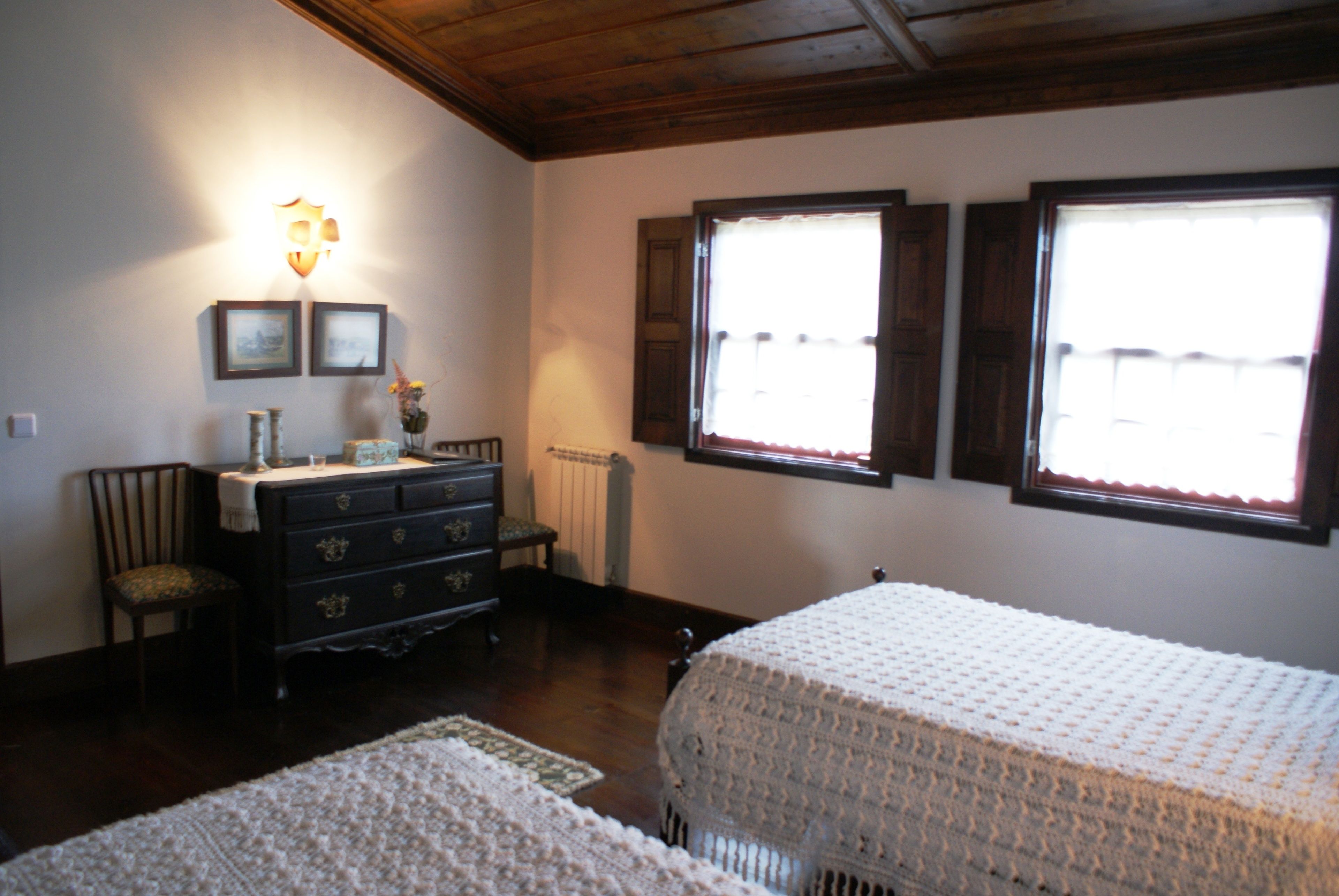 Quarto com duas camas, um penteado, uma cadeira, uma luminária na parede e duas janelas.