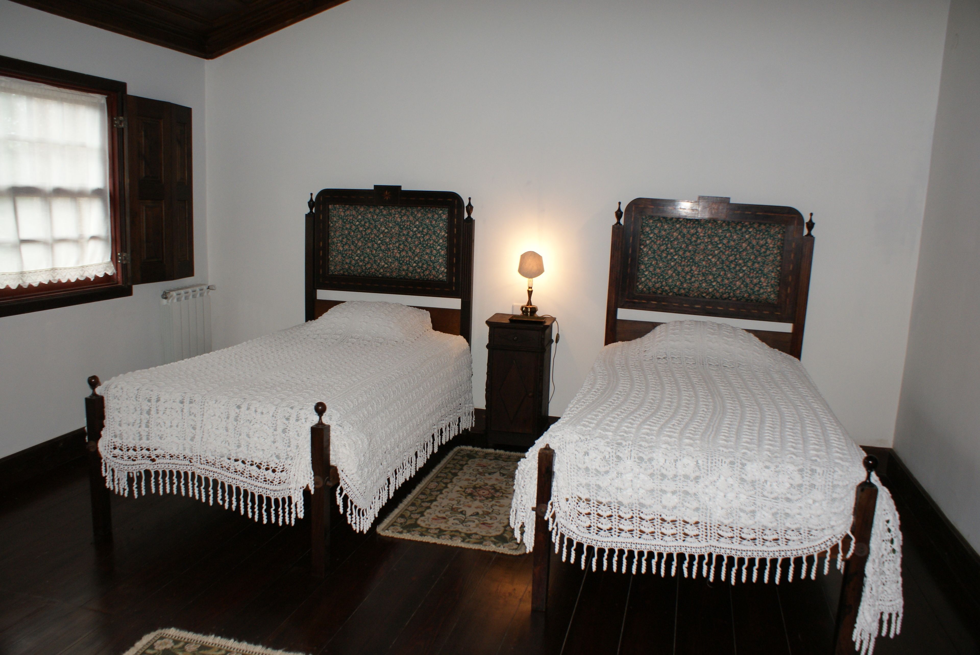 Two single beds with white lace covers, a wooden floor, a small bedside table with a lamp, and a window with a wooden frame.