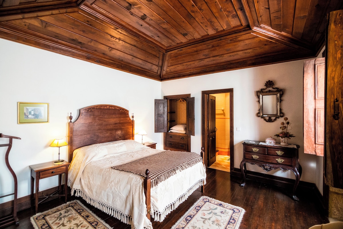 A traditional bedroom with a wooden ceiling, a large bed with a headboard, a nightstand, a dresser, and a mirror.