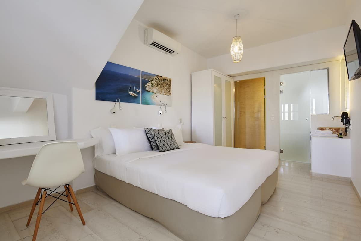 A bedroom with a bed, a desk, and a chair. There is a painting on the wall, a ceiling light, and a door leading to another room.