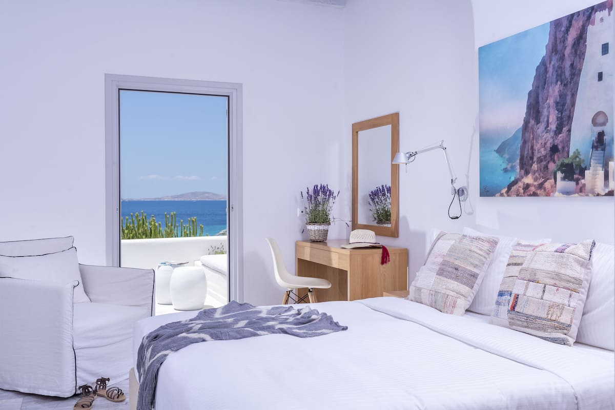A bedroom with a bed, a desk, a chair, and a view of the sea.