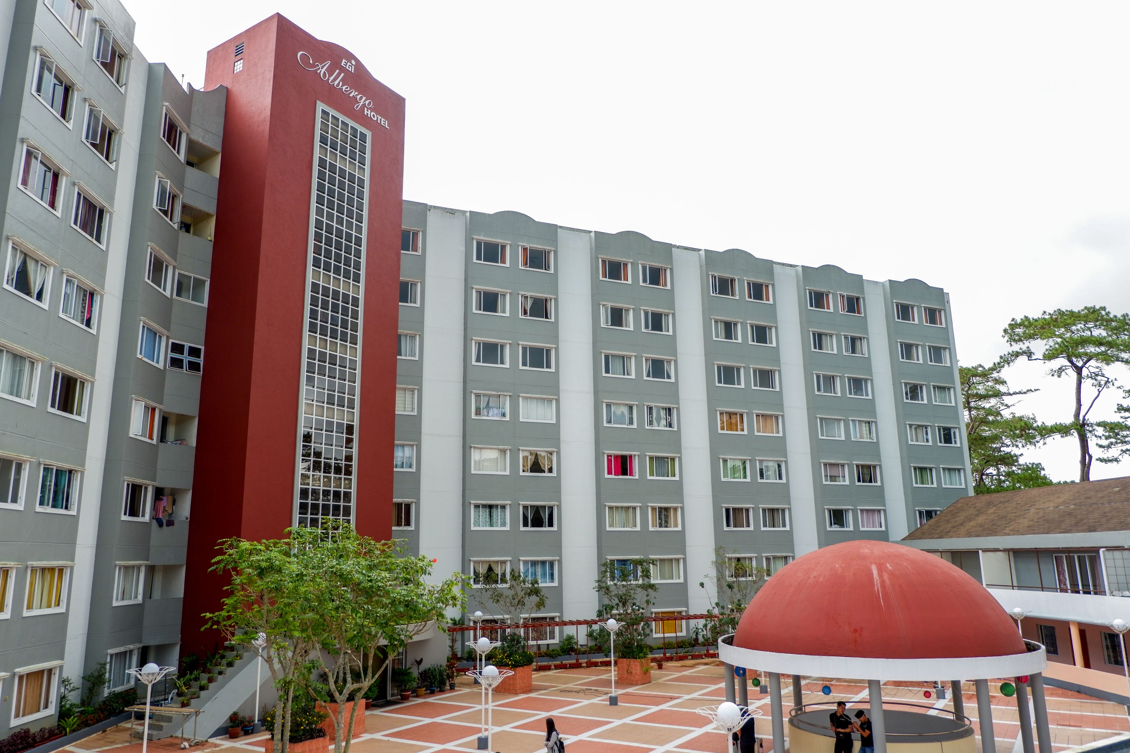 A multi-story hotel with a distinctive red and grey facade, a central staircase, and a covered outdoor area with a dome structure.