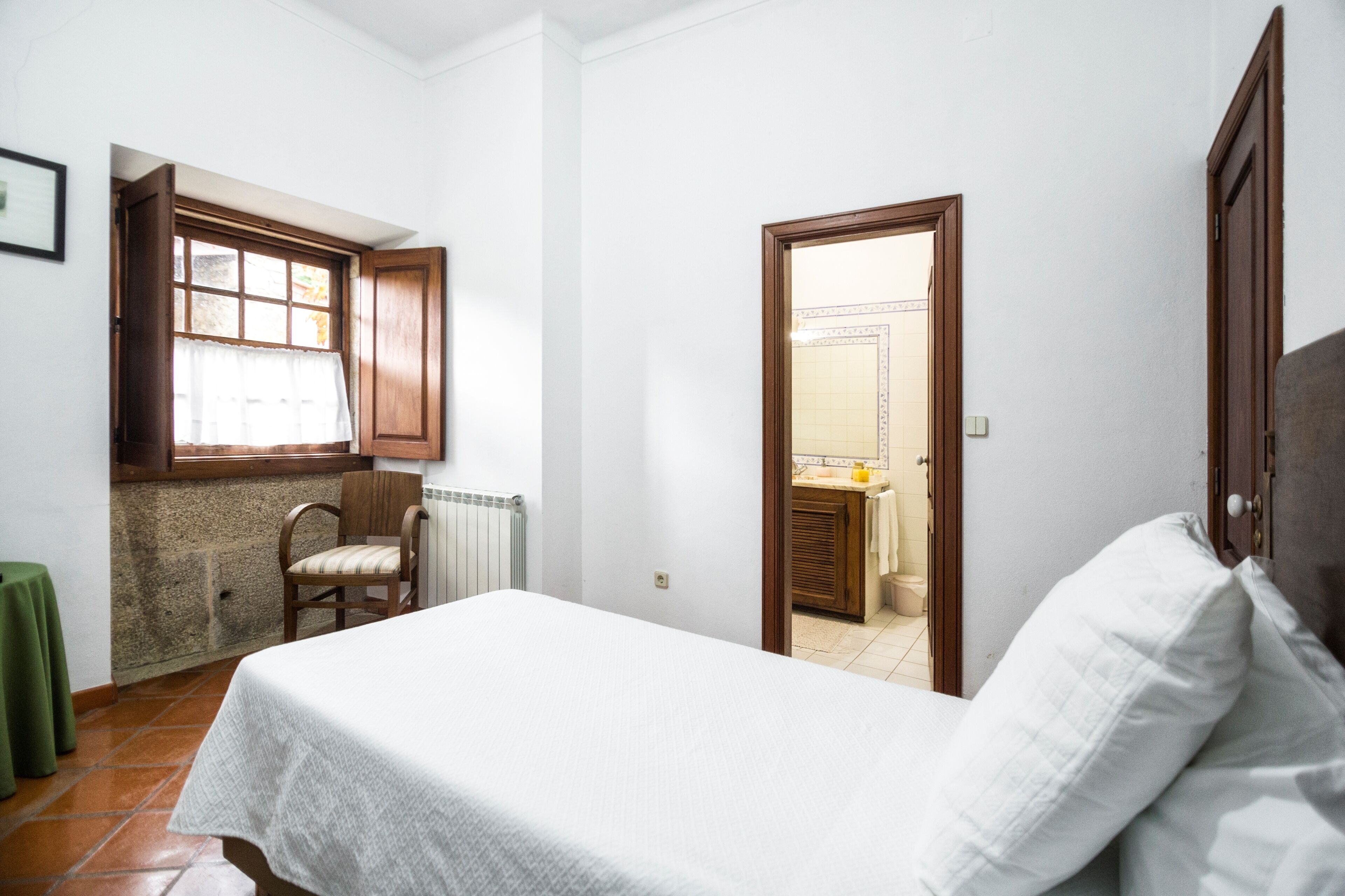 A small, clean room with a bed, a chair, a window with a wooden frame, and a door leading to a bathroom.