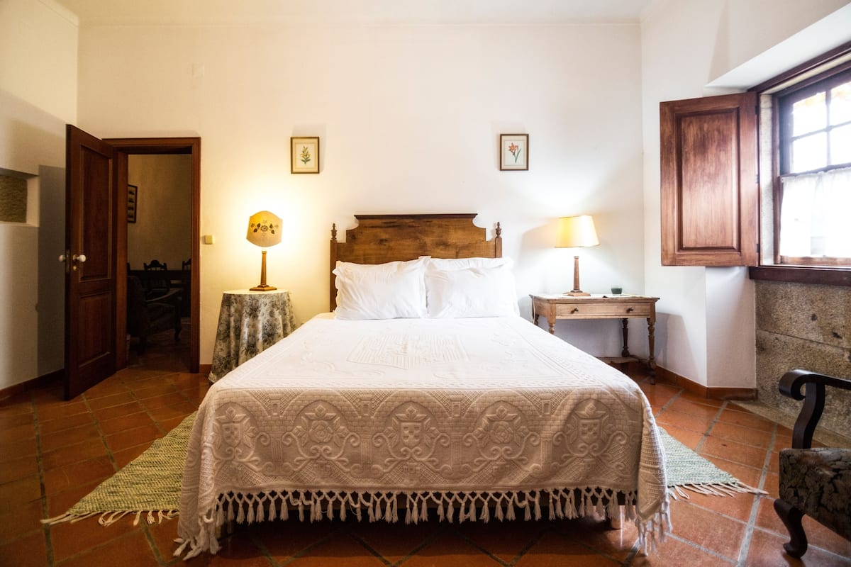 A bedroom with a large bed, two bedside tables with lamps, a wooden headboard, and a window with curtains.
