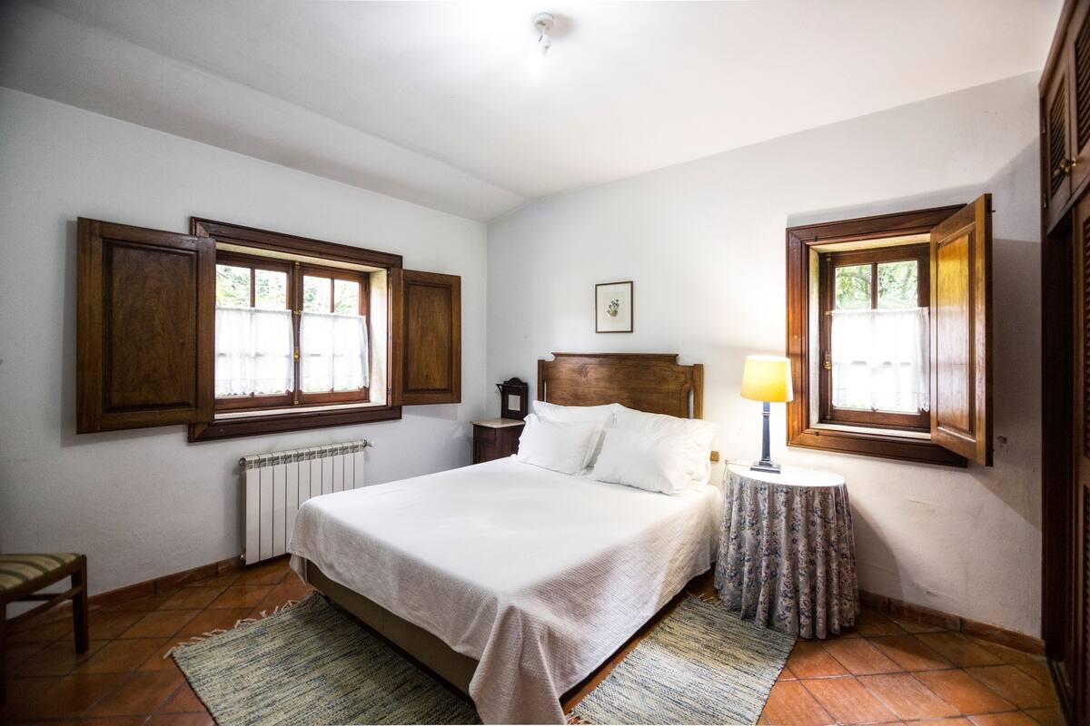 A bedroom with a large bed, two bedside tables, a nightstand, a window with wooden shutters, a radiator, and a small table with a lamp.