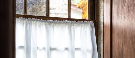 A corner room with a wooden chair, a window with a white curtain, and a stone wall.