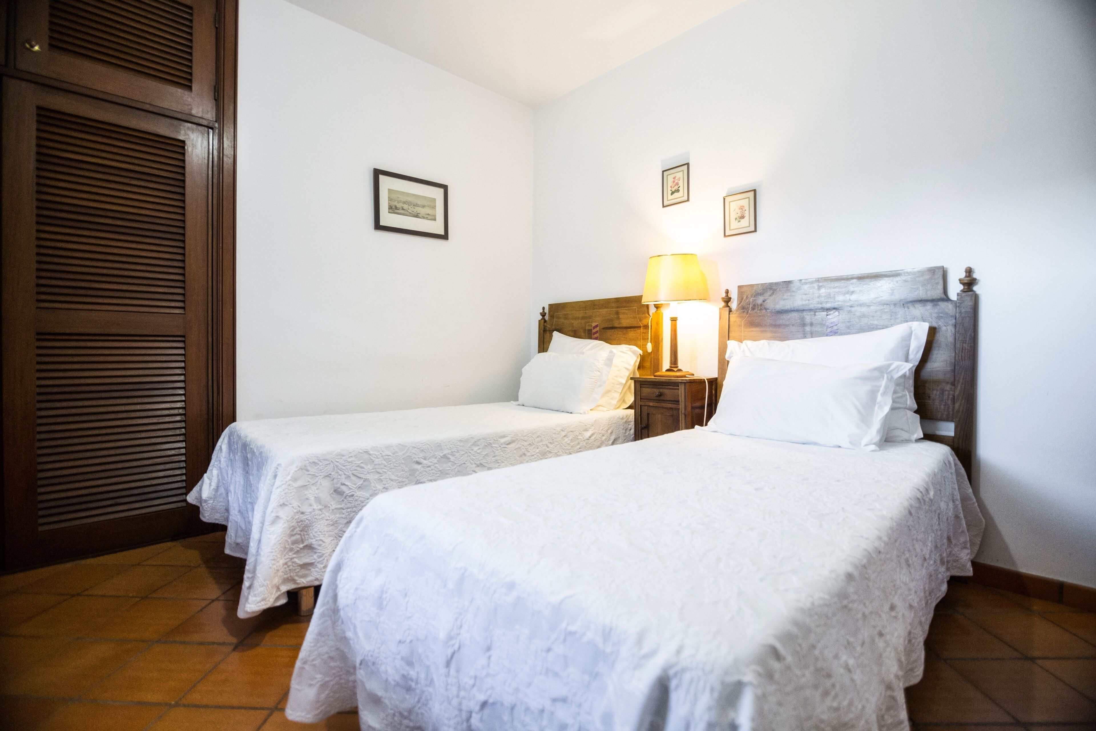 A hotel room with two single beds, a wooden headboard, a nightstand with a lamp, and a door with a wooden shutter.