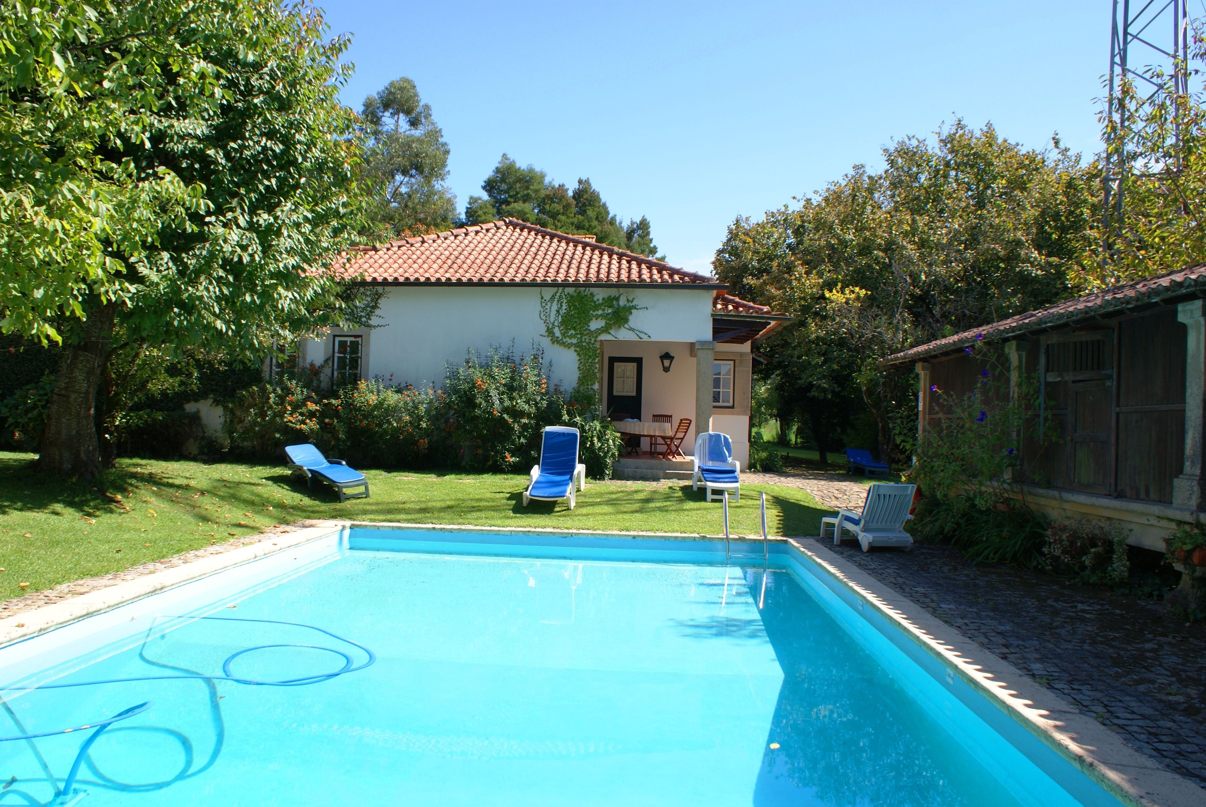 Piscina com espreguiçadeiras, casa com telhado em cerâmica e vegetação ao redor.