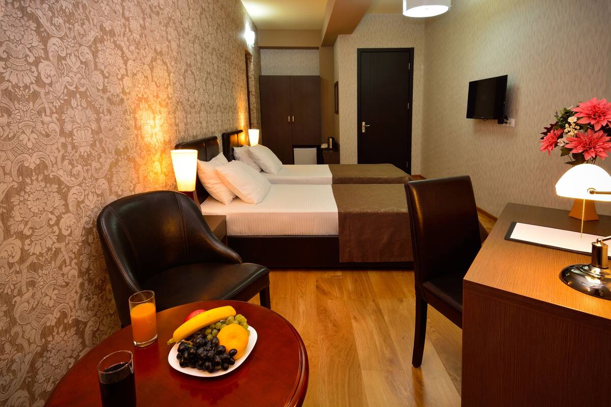 Habitación de hotel con cama, dos sillones, escritorio, televisor y una mesa con fruta y bebidas.