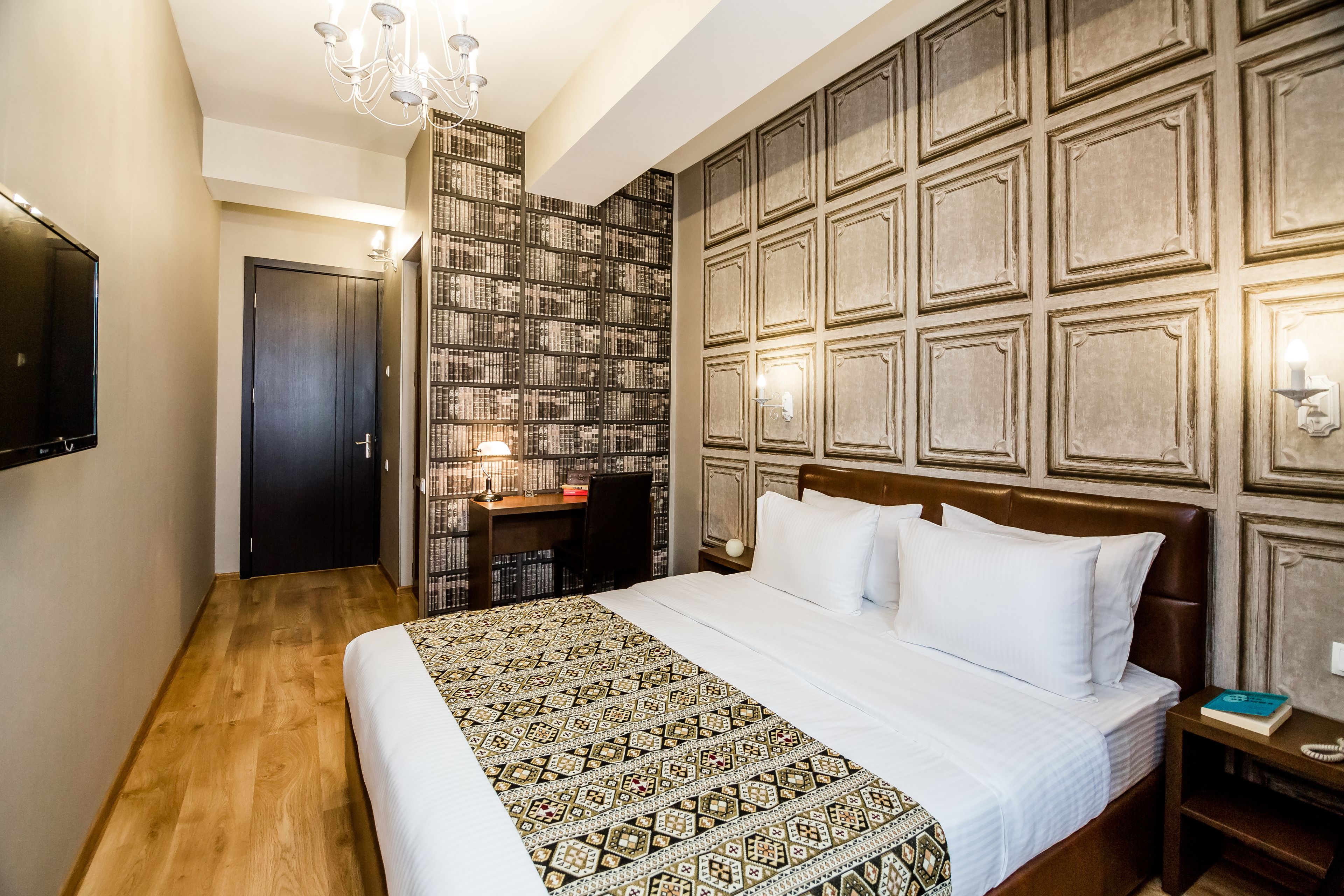 A hotel room with a large bed, a desk with a chair, a bookshelf, and a TV on the wall.