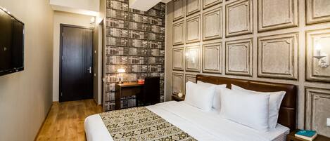 A hotel room with a large bed, a desk with a chair, a bookshelf, and a TV on the wall.