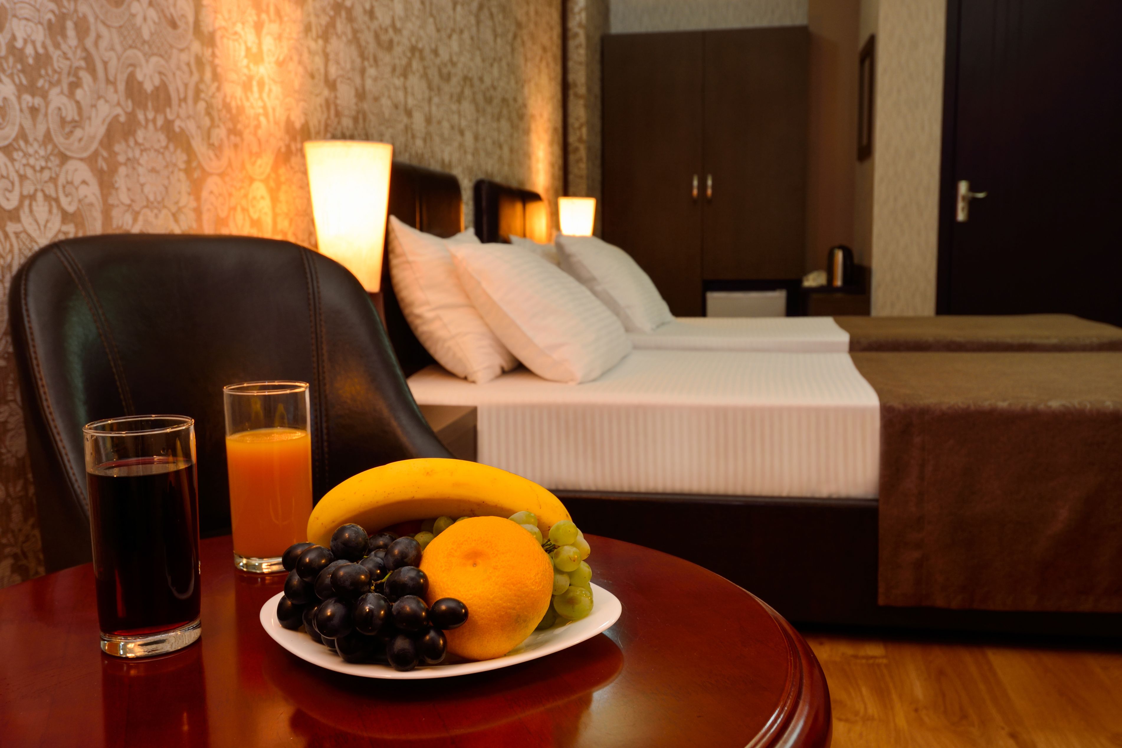A hotel room with a bed, a bedside table with glasses of juice, and a plate of fruit.