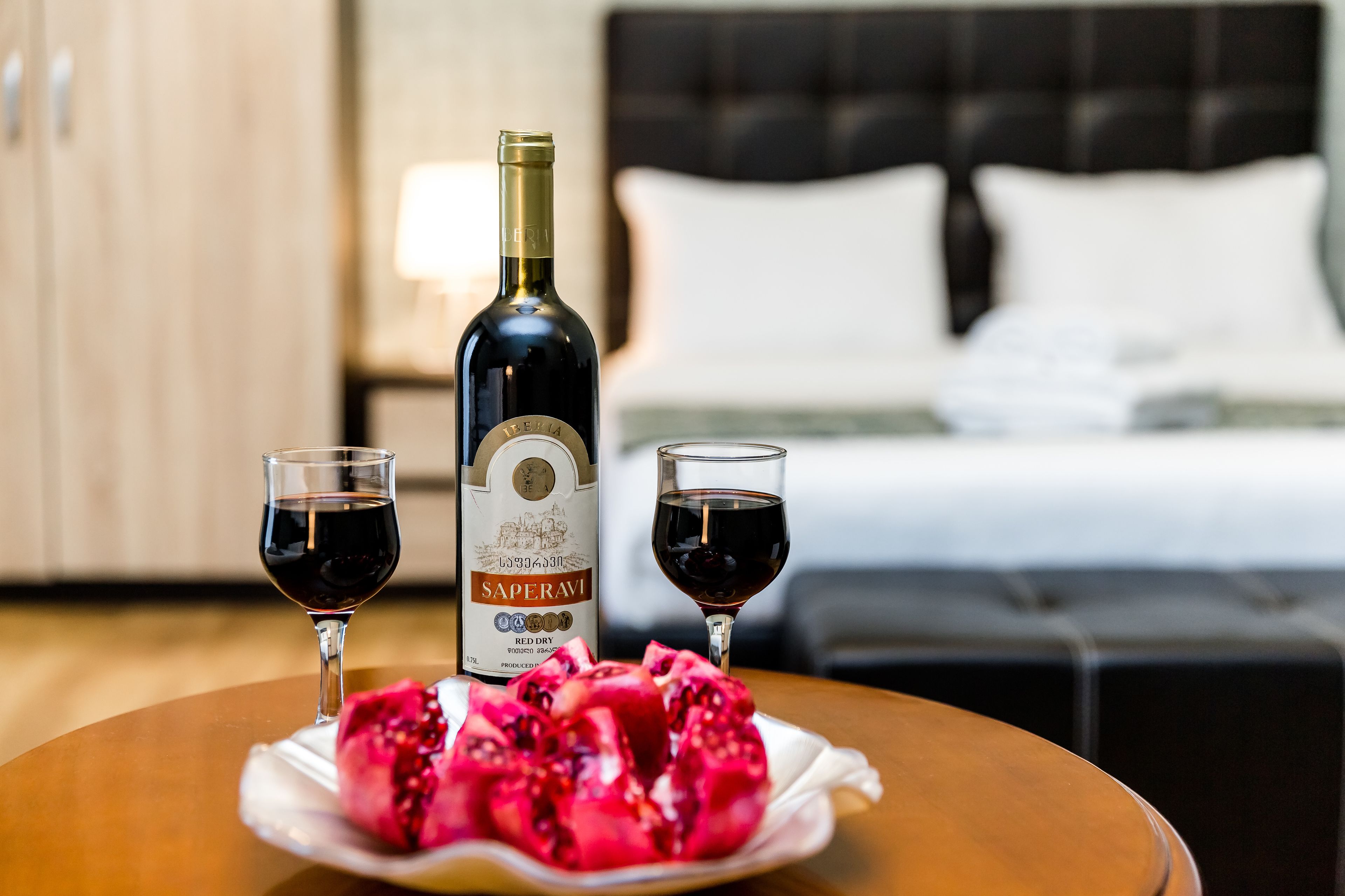 Eine Flasche Saperavi-Wein, zwei Rotweingläser und eine Schale mit Granathomaten auf einem Tisch in einem Hotelzimmer.