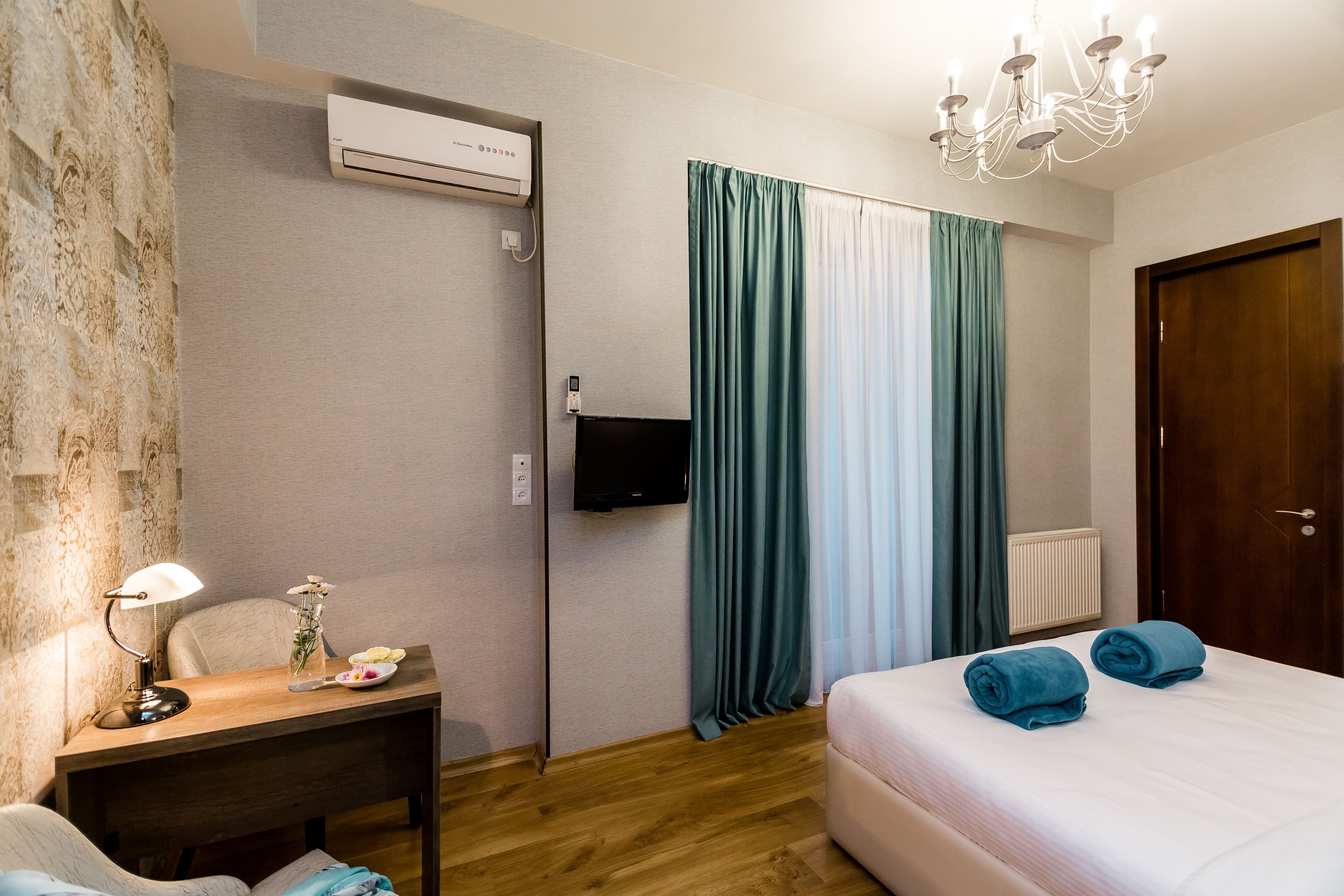A hotel room with a wooden desk, a chair, a bed with blue towels, a TV, and a chandelier.