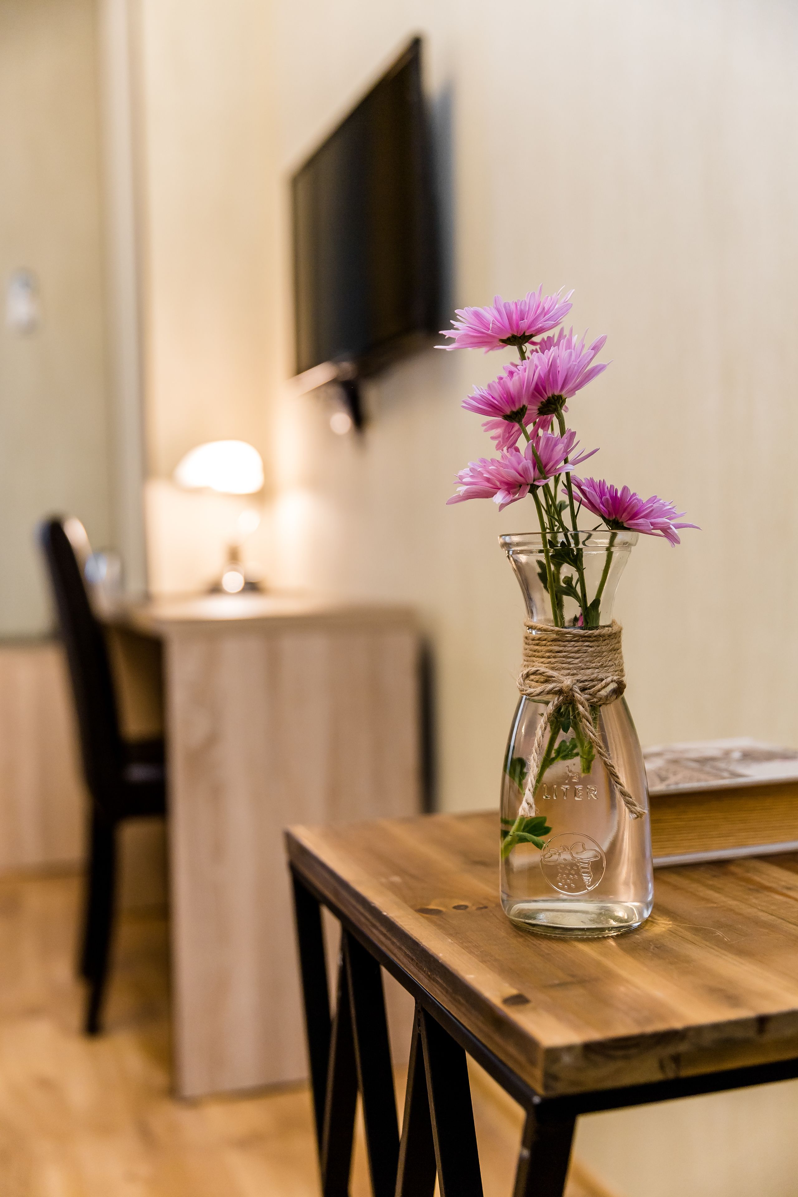 Um vaso de vidro transparente com flores cor-de-rosa sobre uma mesa de madeira num quarto com uma TV fixada na parede.