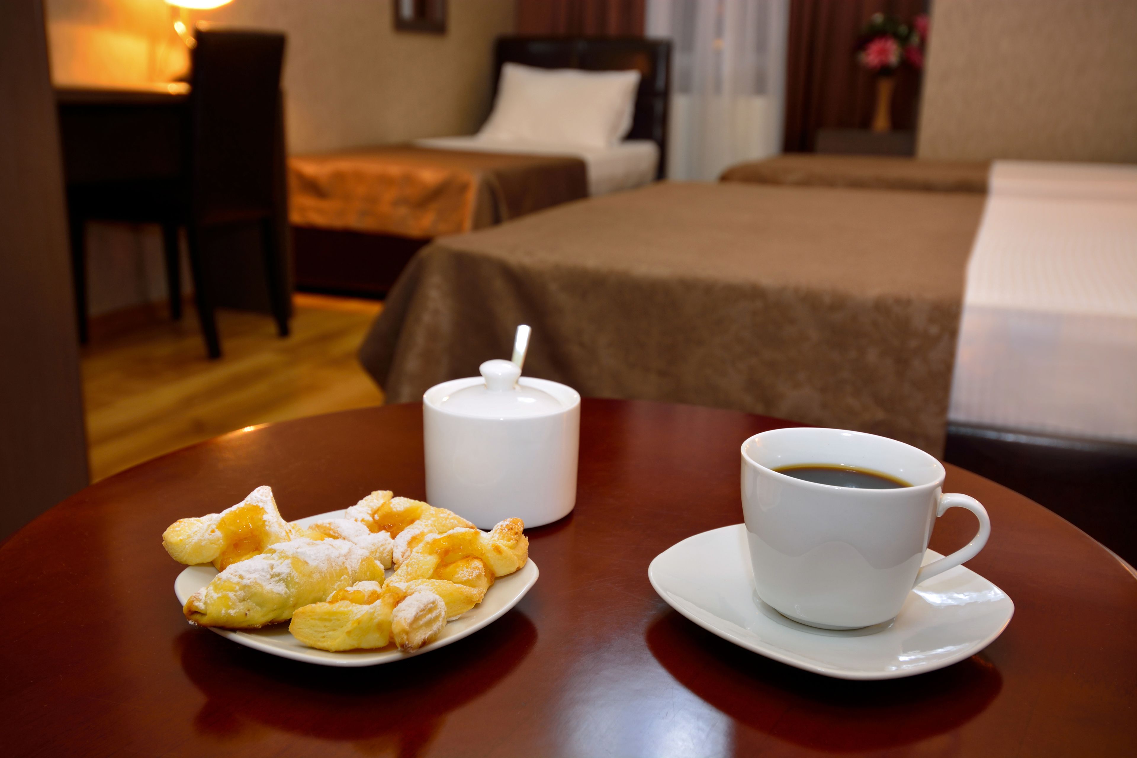 A hotel room with a bed, a bedside table with a cup of coffee and pastries, and a chair.
