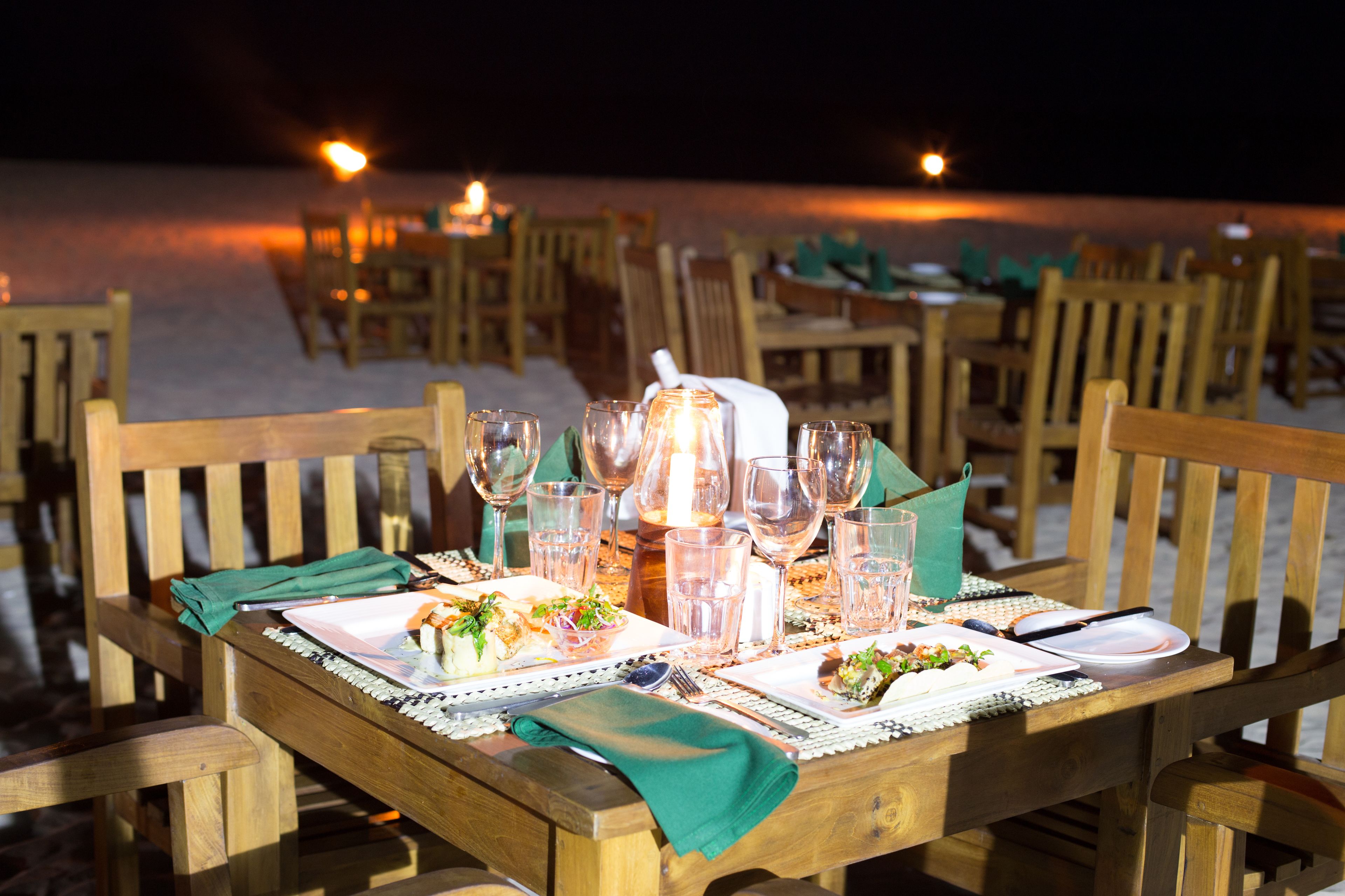 A wooden dining table set with plates, glasses, and cutlery, surrounded by chairs on a beach.