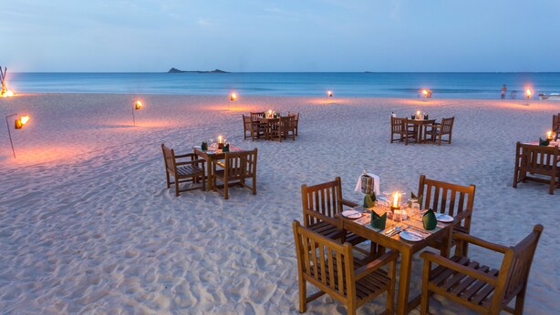 Un espace de restauration situé en bord de mer, avec des tables et des chaises disposées sur le sable, chacune agrémentée de bougies et d’une décoration de table.