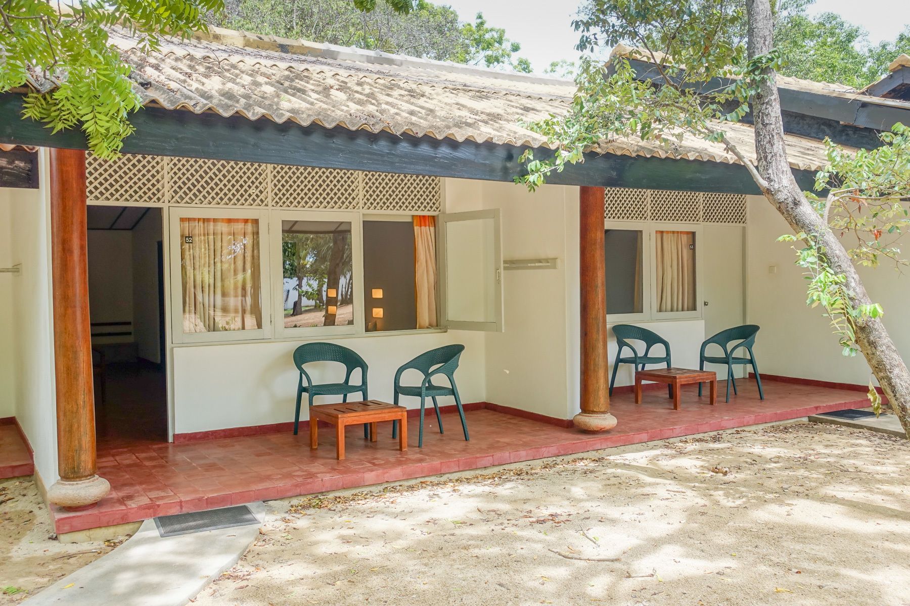 A covered outdoor seating area with two tables and four chairs, a tiled roof, and a view of the surrounding greenery.