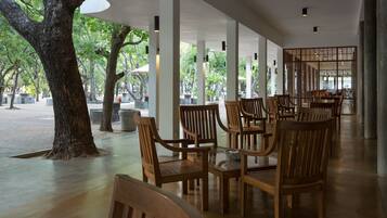 A covered outdoor seating area with wooden tables and chairs, surrounded by trees and a view of the interior.