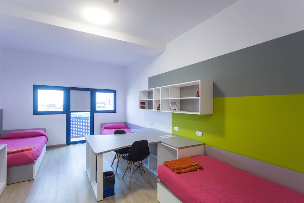Une chambre moderne et compacte, dotée d’un bureau, de deux lits, d’une chaise et d’une fenêtre.
