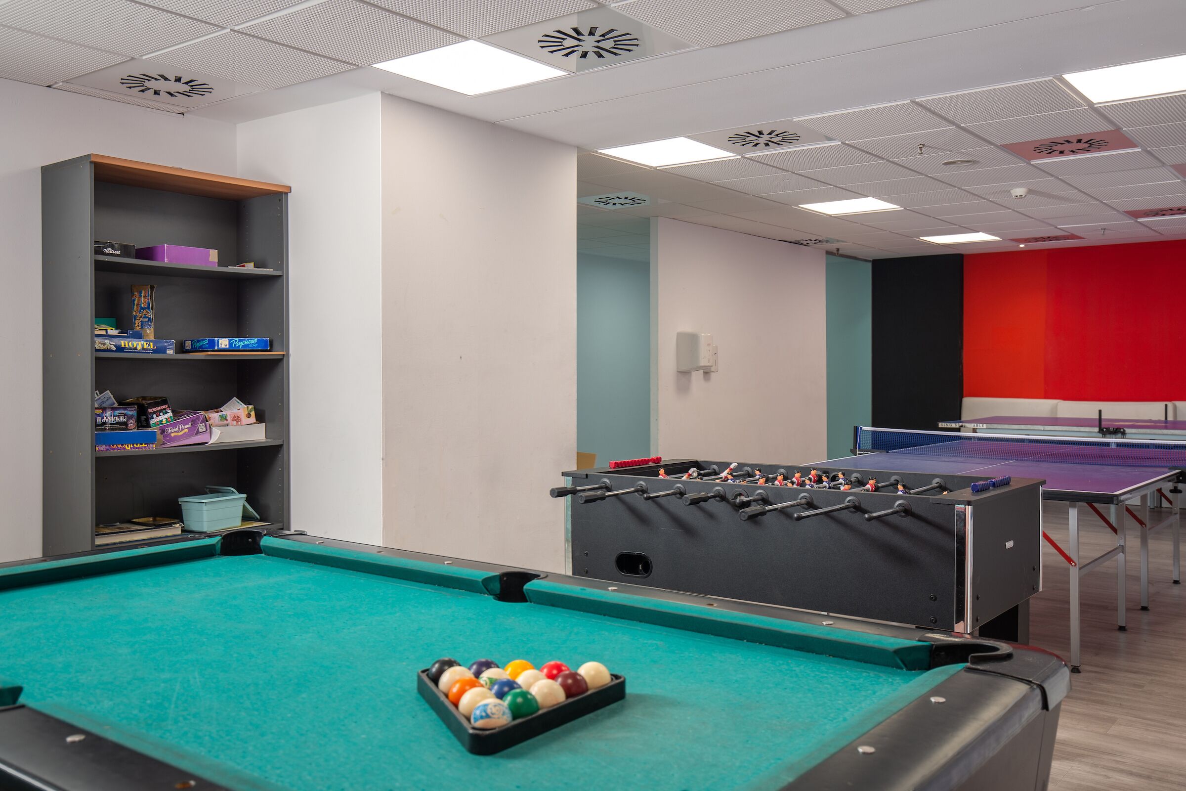 A pool table with colorful balls, a foosball table, and a ping pong table in a room with white walls and a red accent wall.