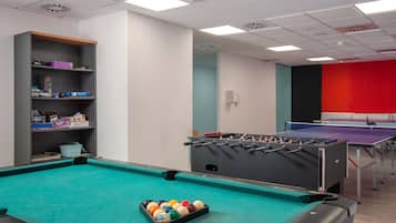 A pool table with colorful balls, a foosball table, and a ping pong table in a room with white walls and a red accent wall.
